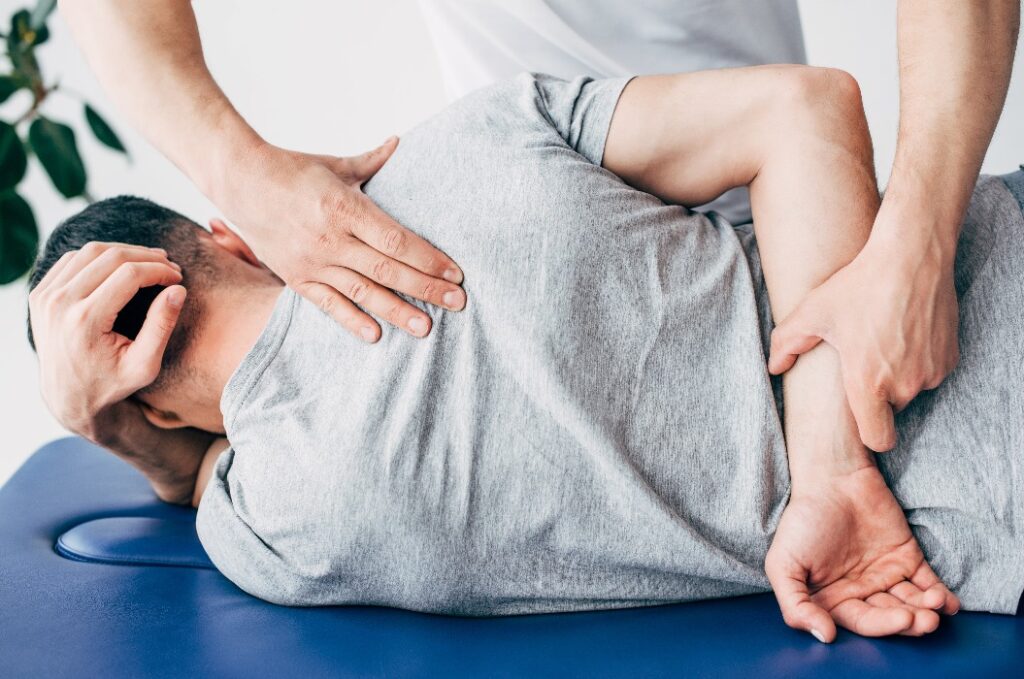 Rückenansicht eines Chiropraktikers, der den Rücken eines Mannes massiert, der auf einem Massagetisch im Krankenhaus liegt